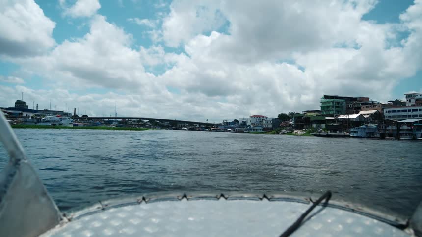 A wide point of view from the bow of a small aluminum boat on a wide river shows a city skyline with buildings crowded up a hill and a concrete bridge spanning the water under a bright blue sky with fluffy white clouds