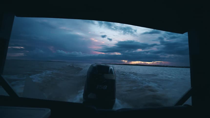 A point of view shot from the back of a dark boat looks out over a churning lake or wide river showing a black outboard motor pushing the vessel forward into a dramatic sunset with dark storm clouds