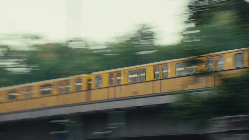 A yellow commuter train speeds across an elevated track from left to right its fast motion blurring the green trees and buildings in the background