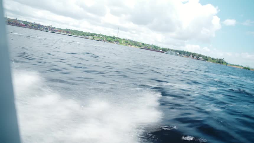 The video shows a lowangle fastmoving shot from a boat traveling across a wide expanse of blue water generating a bright white wakethe shoreline is visible in the background under a partially cloudy sky featuring a distant town or industri