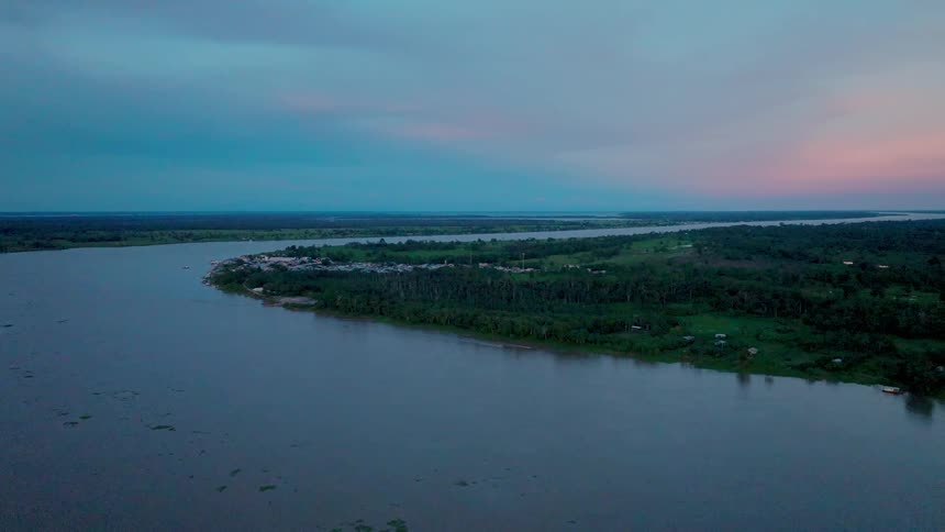 An aerial extreme wide view shows a large wide river winding through a dense green rain forest with a small clustered settlement along the near bank under a dark twilight sky