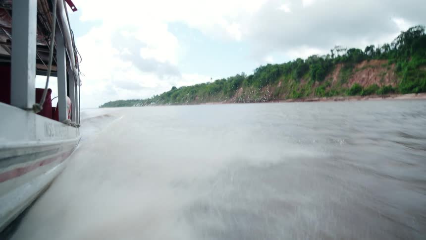 A low angle view from the side of a fast moving boat shows a wake rushing over the wide river with a forested earthen bank in the background under a cloudy sky
