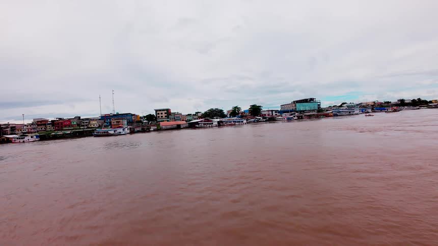 A wide brown body of water flows toward the camera past a crowded shore lined with lowrise buildings and docked watercraft under a pale cloudcovered sky