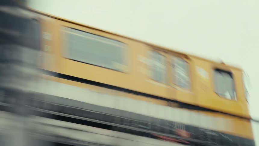 Slow shutter shot of a long yellow transit vehicle that moves quickly along a raised railway structure a dark fence parallels the track and green leaves appear in the corner against an overcast sky