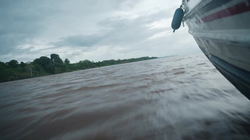 A close low angle view shows the side of a boat moving quickly over the wide choppy brown water of a river with a green rain forest shoreline visible under a cloudy sky