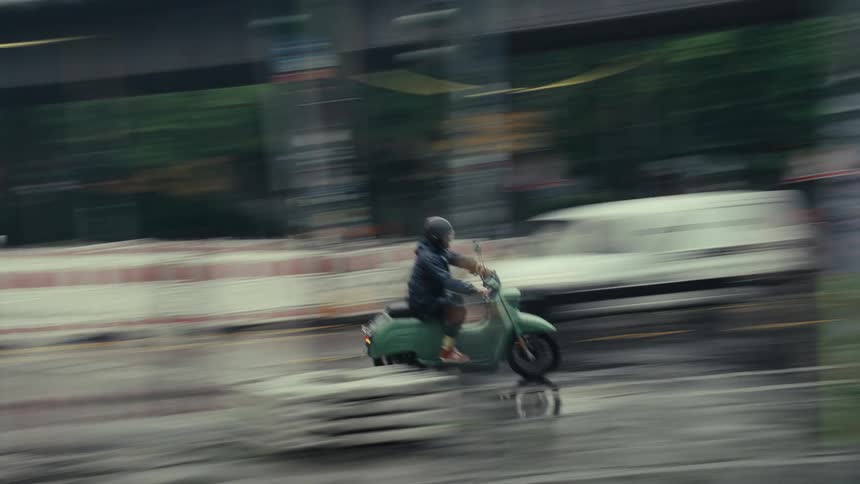 A man on a green scooter speeds through a rainy city street captured with strong motion blur - slow shutter