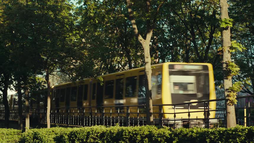 A fast moving bright yellow subway train rushes past a black metal fence and lush green park trees on a bright and sunny afternoon