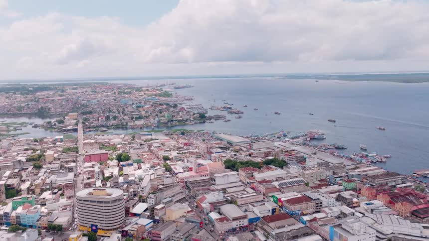 An aerial extreme wide view shows a dense coastal city with low colorful buildings a wide river estuary and many boats near a busy port under a cloudy sky