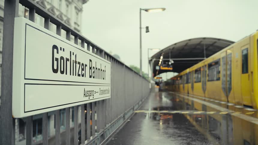 A yellow subway train arrives at the goerlitzer bahnhof platform on a rainy day with the station sign in the foreground and the wet platform glistening under the arched roof