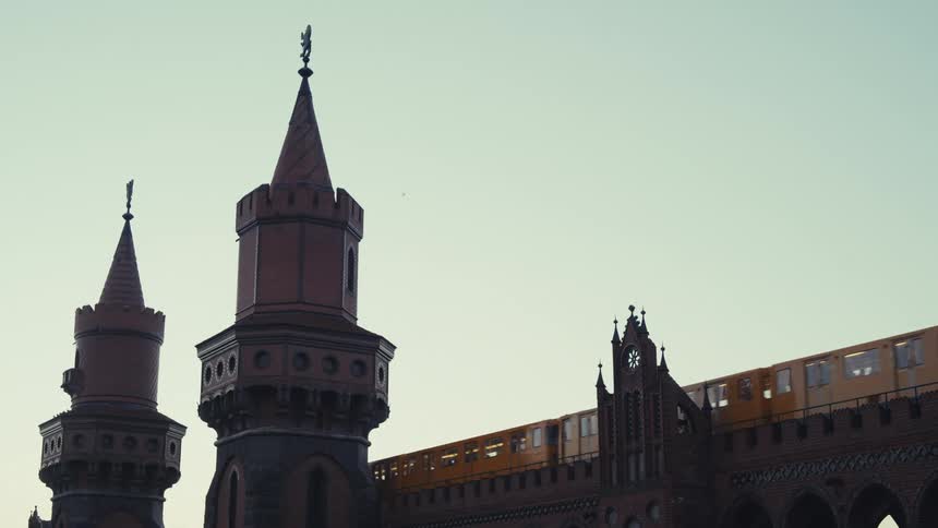 A yellow train moves across the upper level of a large red brick bridge structure on the Oberbaum bridge in Berlin, passing between gothicstyle towers under a light sky