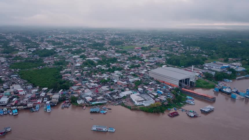 An aerial view shows a wide murky river next to a large town clustered buildings line the shore a sizable structure with a metal roof stands near the water and numerous watercraft are moored along the bank under a gray sky