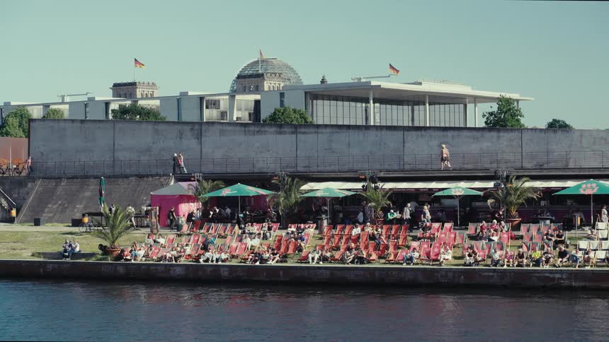 Numerous people sit in reclining chairs on a riverbank protected by green umbrellas and small trees facing a calm waterway below a large concrete embankment supporting a complex of white buildings with dome features and flying flags