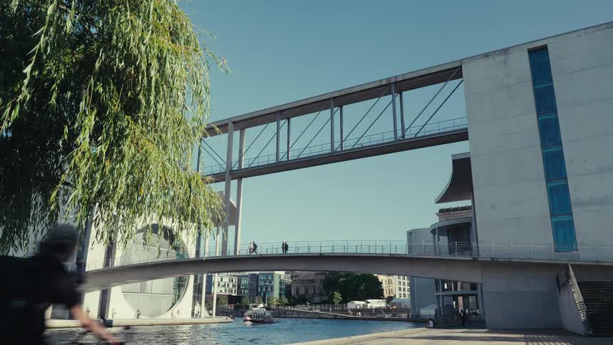 The video shows an architecturally unique building complex with two large flattopped concrete wings connected by a highlevel enclosed bridge and a lower arched pedestrian bridge spanning a canal two people are visible walking across the lower ar