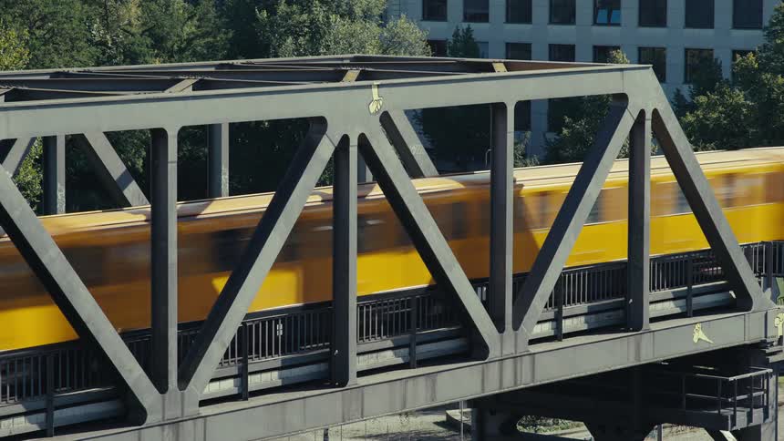 A high angle medium view shows a blurry yellow subway train quickly passing over a gray truss bridge on an elevated track above a treelined city street (slow shutter)