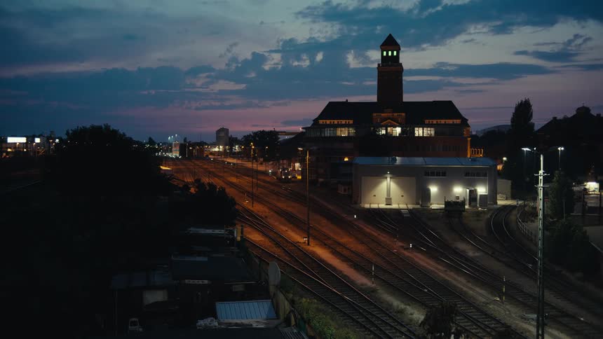 A camera shot shows a wide view of numerous railroad tracks leading toward a large brick building with a tower illuminated by track lighting and streetlights during the transition from sunset to night