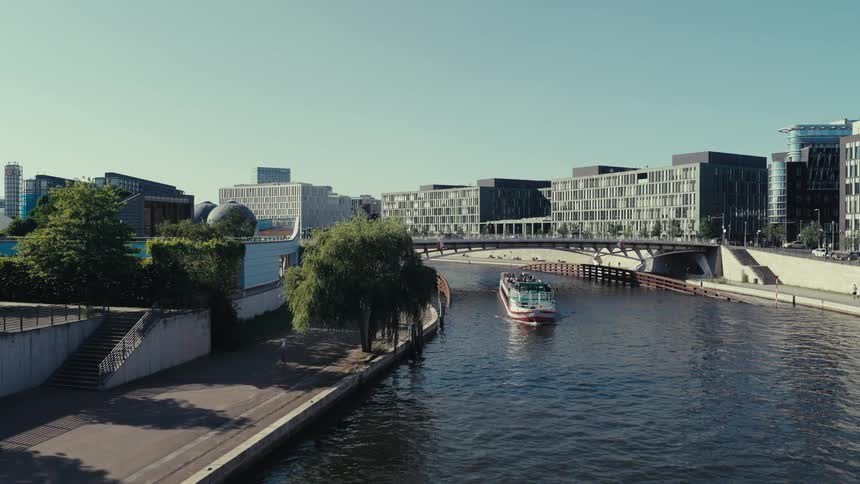 A jogger runs along the side of a canal where a ferry boat is moving on the water under a bridge