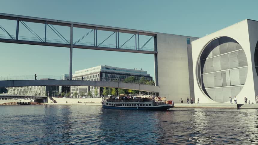 A long wide view of the river spree in berlin shows a tour boat passing under the connecting bridges between the modern paul lobe haus and the marie elisabeth lueders haus on a bright sunny day