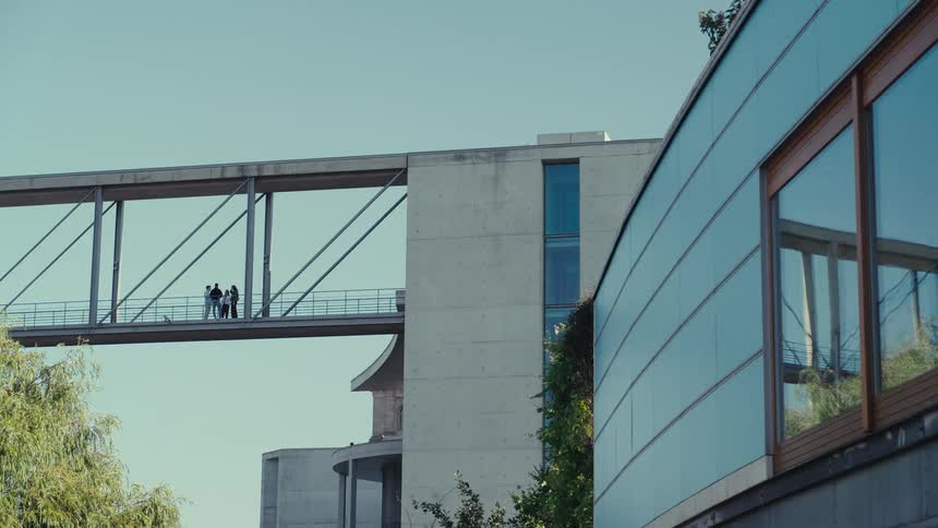 Two structures connect with an elevated walkway several figures stand on the overhead passage a glass facade with wooden frames sits in the right foreground and green tree tops line the lower edge of the frame under a clear atmosphere