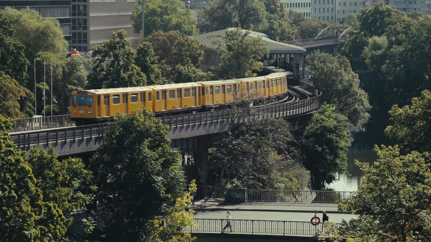 A yellow elevated train leaves a station and moves across a bridge surrounded by lush green trees with modern city buildings visible in the background and pedestrians walking below