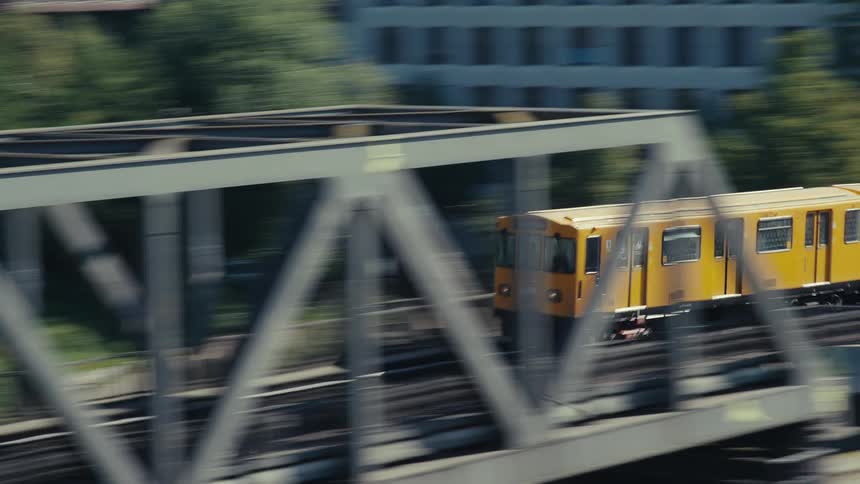 Slow shutter tracking shot of a fast yellow subway train rounding a curve on an elevated steel truss bridge surrounded by bright green trees and urban buildings