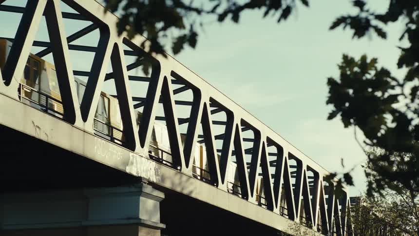 A yellow train crosses a steel truss bridge sun glinting through the lattice and leaves in the foreground