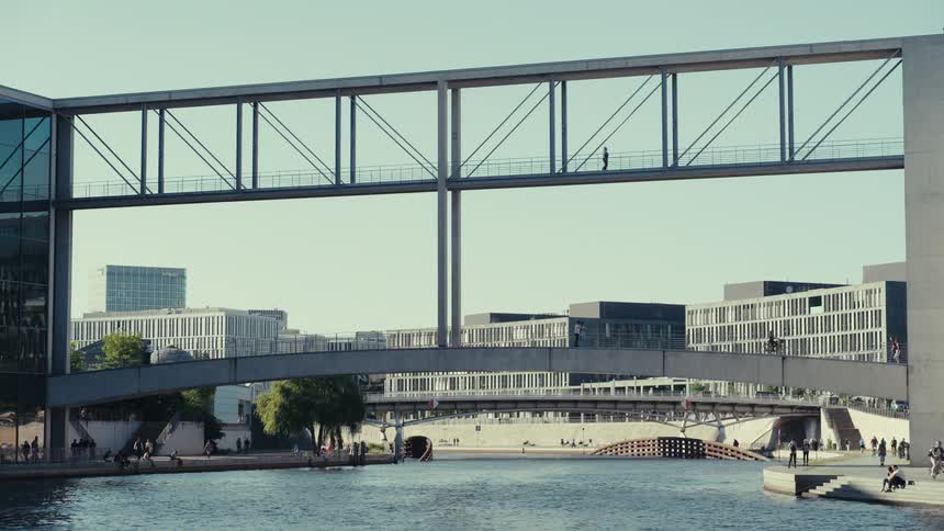 A medium wide view of the marie elisabeth lders haus in berlin shows the two glass and steel pedestrian bridges connecting the political buildings across the river spree with a few people crossing in the distance