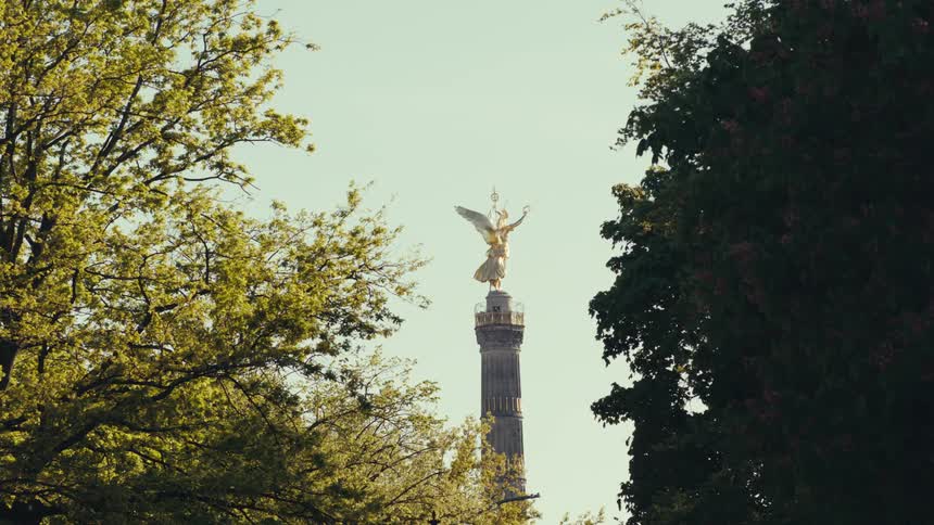 A medium view of the golden victory column statue in berlin rising above the bright green trees framed by dark foliage against a clear sky