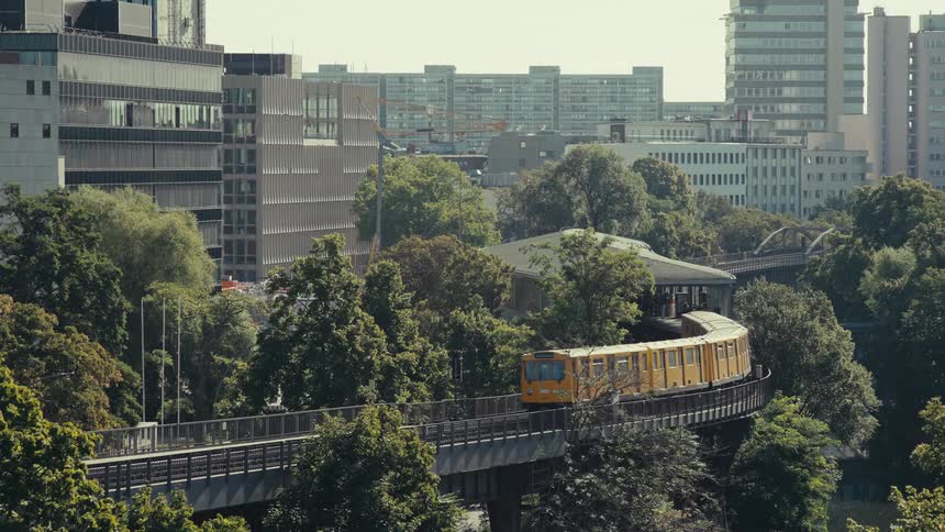 The video is a highangle shot of a yellow train passing over an elevated track that curves through a dense city environmentthe foreground is filled with lush green trees contrasting with the modern tall glass and concrete buildings in the ba
