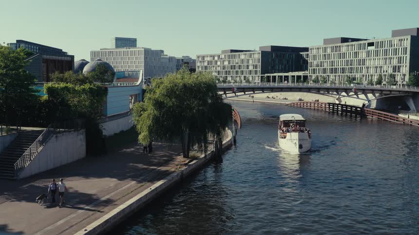 An eyelevel shot captures a white tourist boat moving slowly down a river that flows between modern grey buildings on a sunny day on the shore to the left people are walking and cycling on a path lined with lush green trees