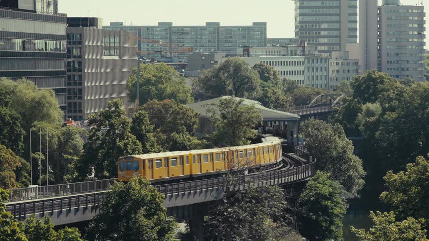A yellow subway train exits an elevated station and moves along a curved track through dense green trees in the foreground with a dense cityscape of modern buildings rising in the background