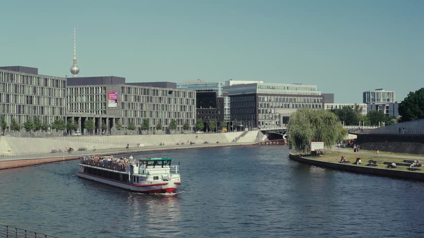 A tour boat with several people on top sailing on a river in the middle of a city