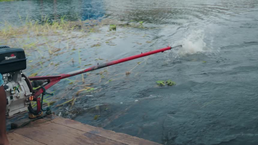 From the back of a wooden boat a longtail motor propeller is lifted out of the water splashing before coming to a stop as the boat glides toward a grassy riverbank