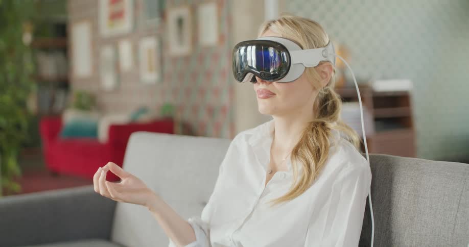 The video is a medium closeup of a blonde woman wearing a white shirt sitting on a couch and utilizing a futuristiclooking white virtual reality vr or meditation headsetshe is seen sitting with her eyes closed and hands slightly raised and