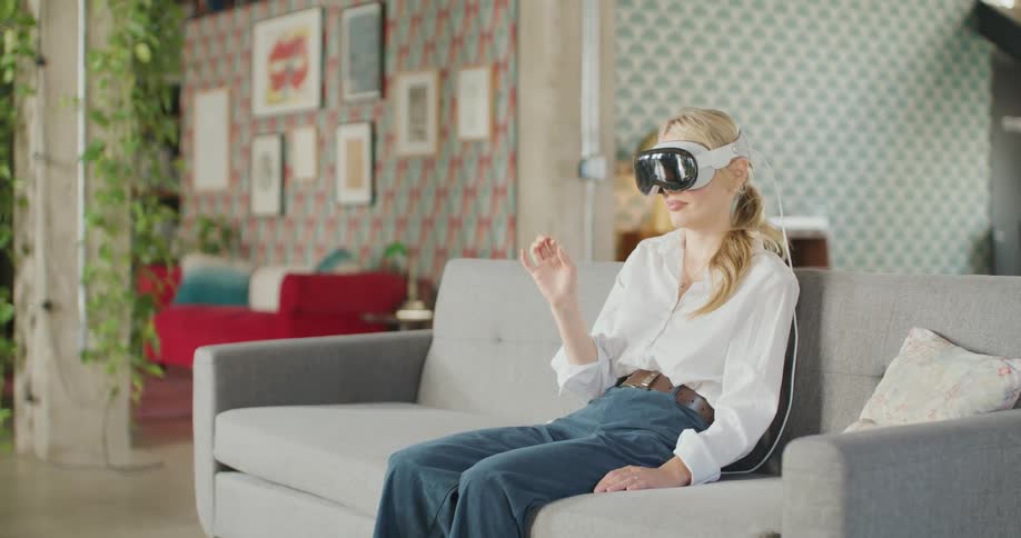 A young woman wearing a white shirt and blue highwaisted trousers is seated on a gray sofa actively using a white virtual reality vr or mixed reality headset she is indoors in a brightly lit modern space with a patterned wallpaper and framed