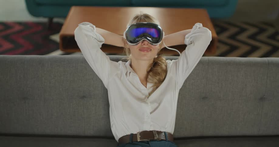 A blonde woman wearing a white shirt and oversized black vr goggles is sitting upright on a gray couch relaxing with her hands clasped behind her head her expression suggests she is enjoying the experience possibly an immersive meditation or re