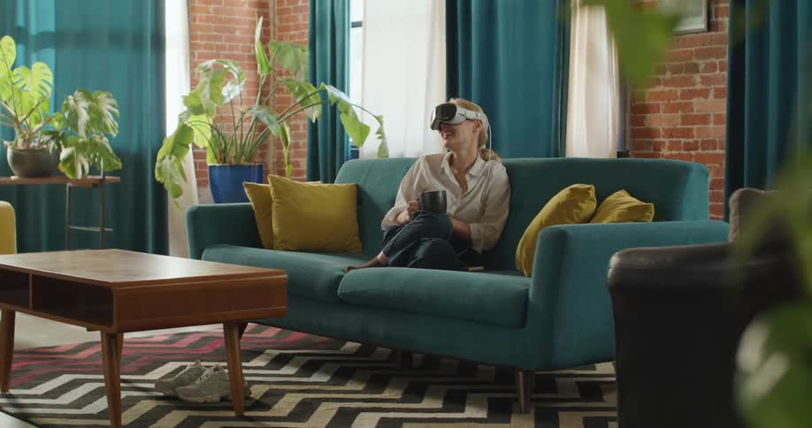 A woman wearing a vr headset and holding a mug sits relaxed on a teal couch looking around and smiling in a brightly lit stylish living room with exposed brick large plants and a geometric rug