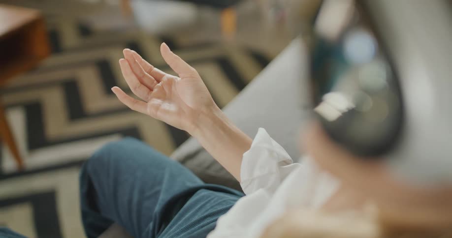 A woman wearing a virtual headset and blue pants sits inside a room raises one hand into the air and slowly opens and closes fingers