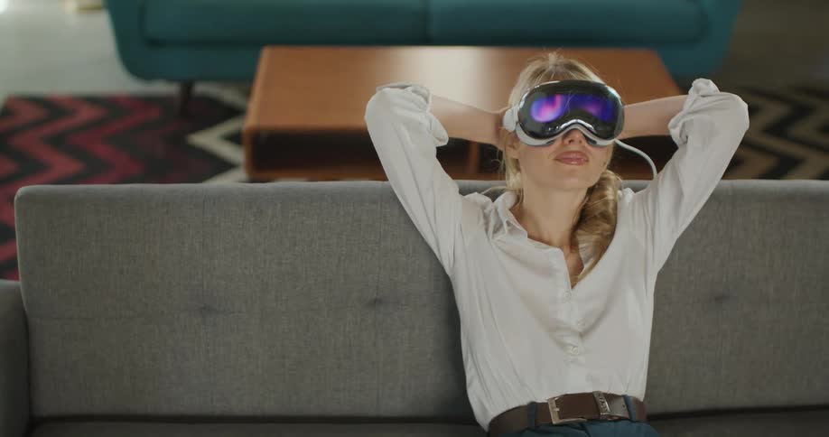 A young woman sits on a sofa wearing an electronic visor over the eyes stretches both arms above the head the indoor room contains furniture items and the area is lit by interior light