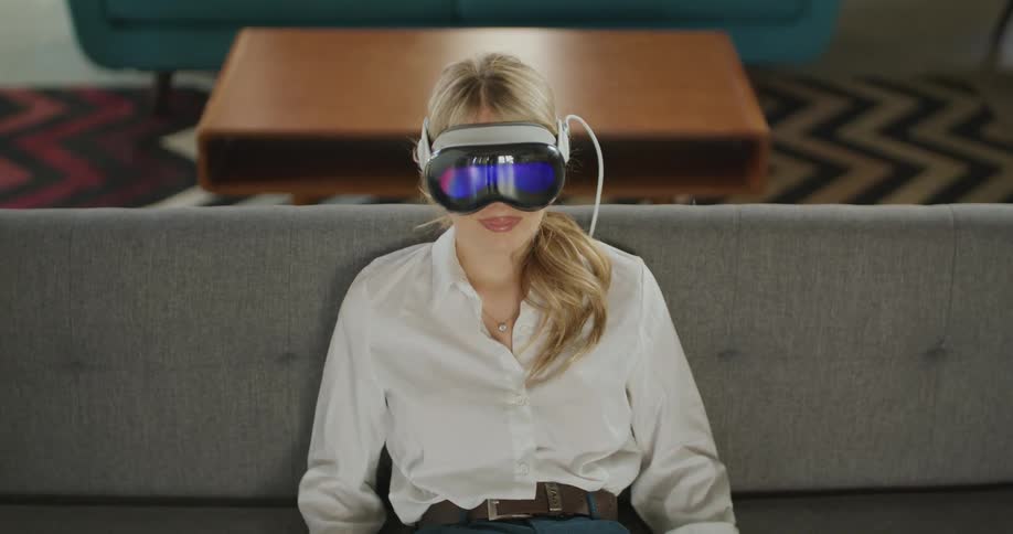 A woman sitting on a couch wearing a vr device then moves the head and looks around raises both arms and places the hands behind the head and the living room contains other household items