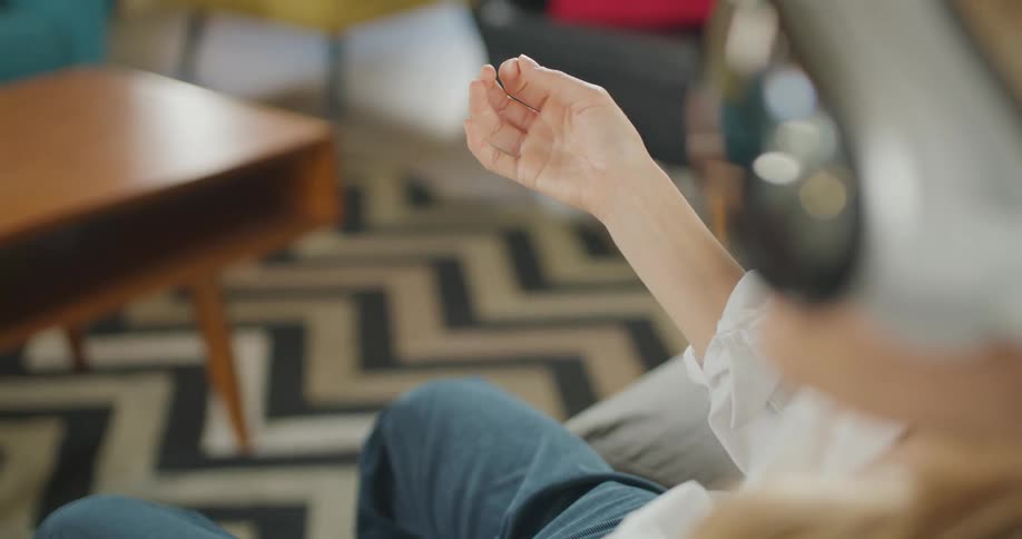 A close view of a person wearing a virtual reality headset and jeans sitting on a sofa reaching out their hand to interact with the air in a brightly lit living room