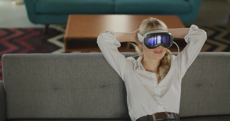 A blonde woman sitting on a couch with her hands on her head wearing a virtual reality headset