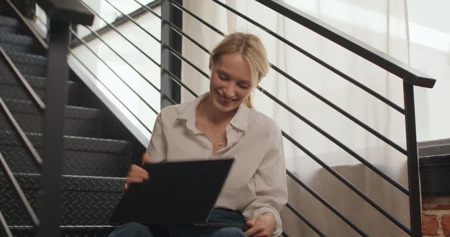 A young blonde woman sitting on a staircase with a laptop on her lap talking to someone and waving during a video call