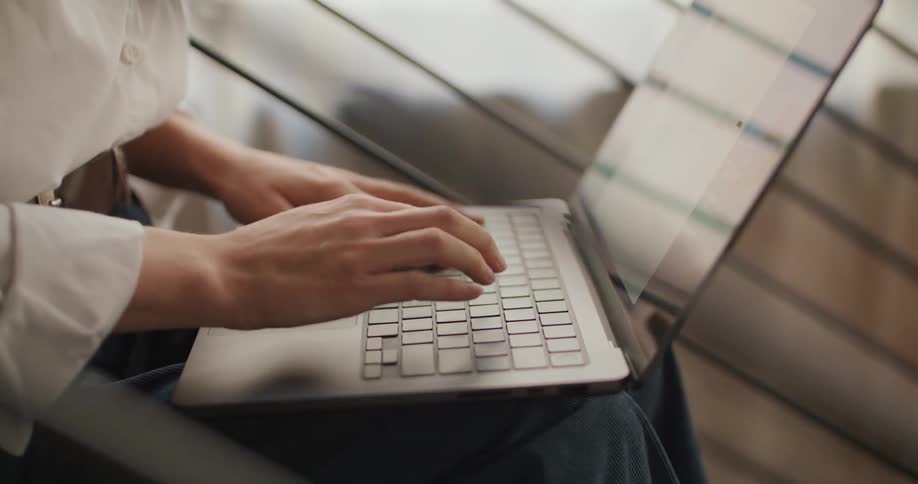 A young woman in a white blouse focuses intently as she types on a silver laptop resting on her lap in a brightly lit space