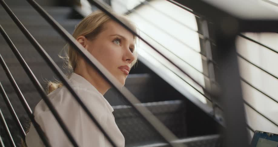 The video is a medium closeup of a blonde woman sitting on a metal staircase in a welllit industrial or modern setting wearing a white collared shirt she is looking down and to the side with a pensive or concerned expression framed by th