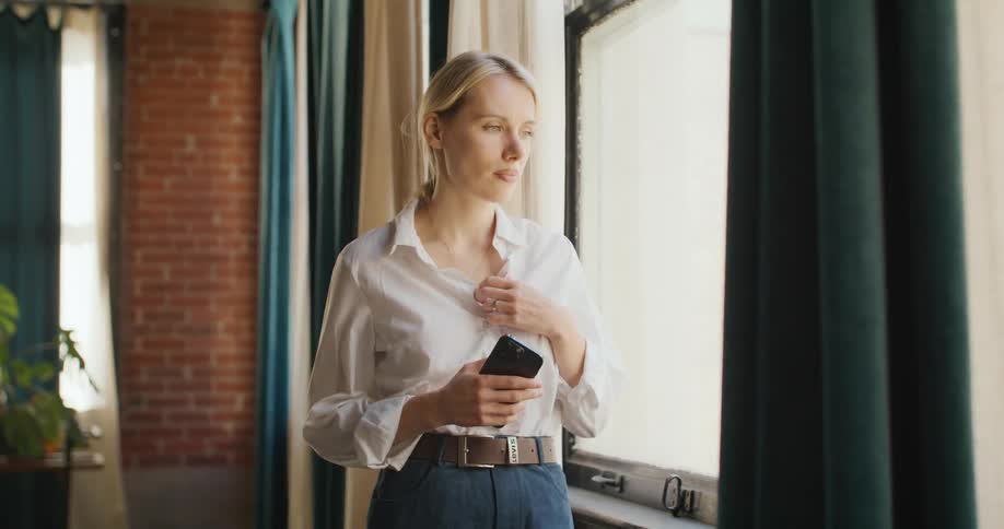 Young woman stands by the window holding her phone