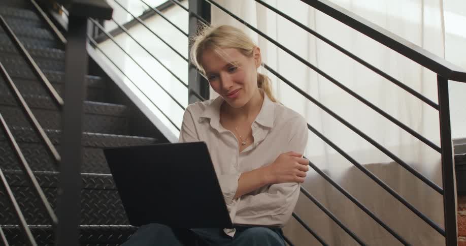 Woman sits on metal staircase smiling at laptop during a video call