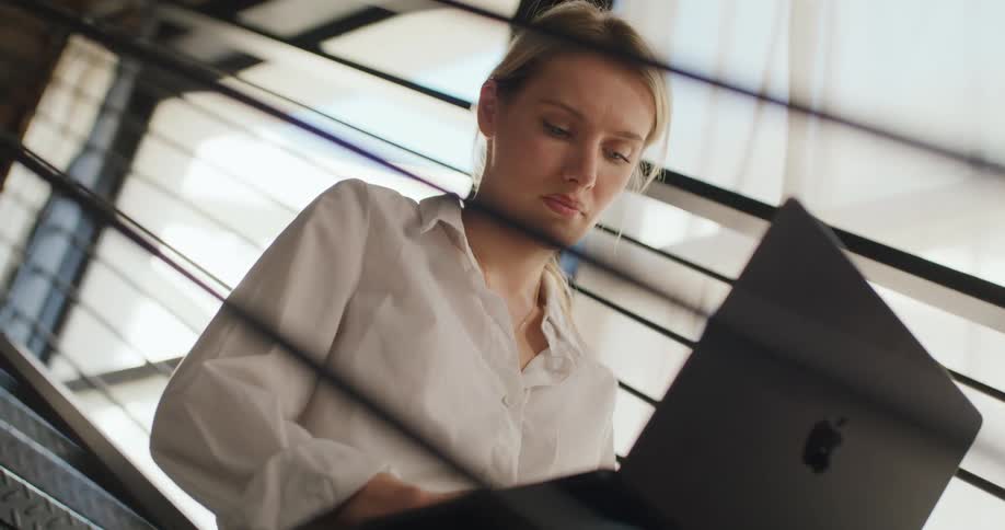 A young woman in a white shirt types on her laptop while sitting on a public staircase at midday
