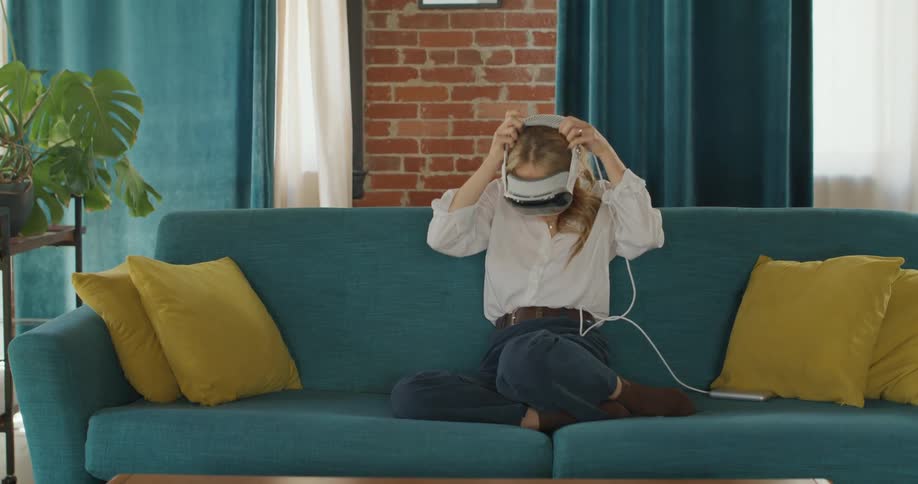 The video shows a woman standing in a living room reaching down to pick up a vr headset and a smartphone from a low wooden table the background features tealblue curtains and a matching sofa with yellow cushions suggesting a modern and stylize