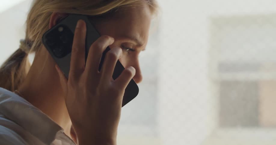 A female is talking on the phone and seems to be annoyed at times while she stands indoors near a window