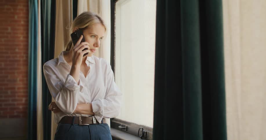 The video is a medium shot of a blonde woman in a white shirt and dark skirt standing and looking at her phone near a windowshe is framed by a bright window with dark green curtains and appears focused or slightly serious while scrolling